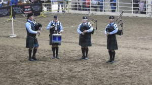 Four people playing bagpipes wearing traditional Scottish clothing on the floor of the National Western Stock Show